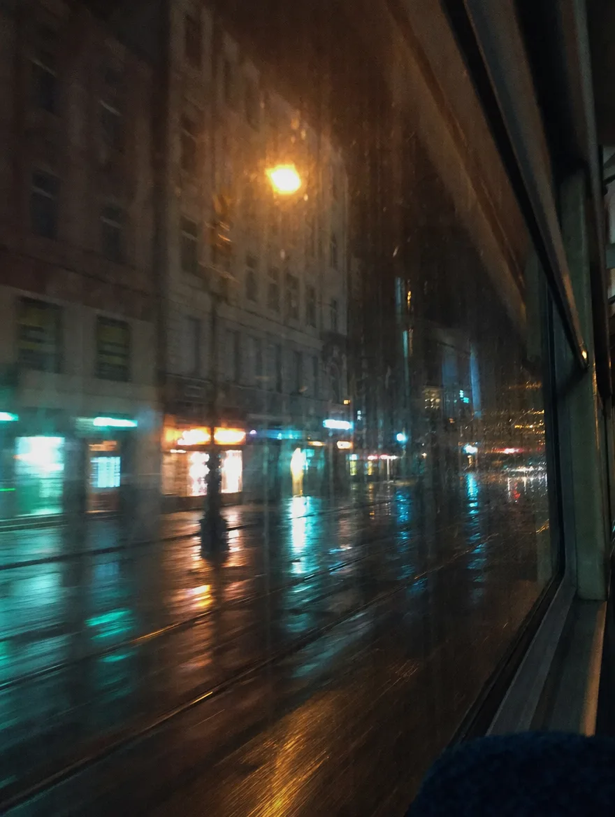 Neony rozmyte w smugach na mokrej szybie tramwaju.