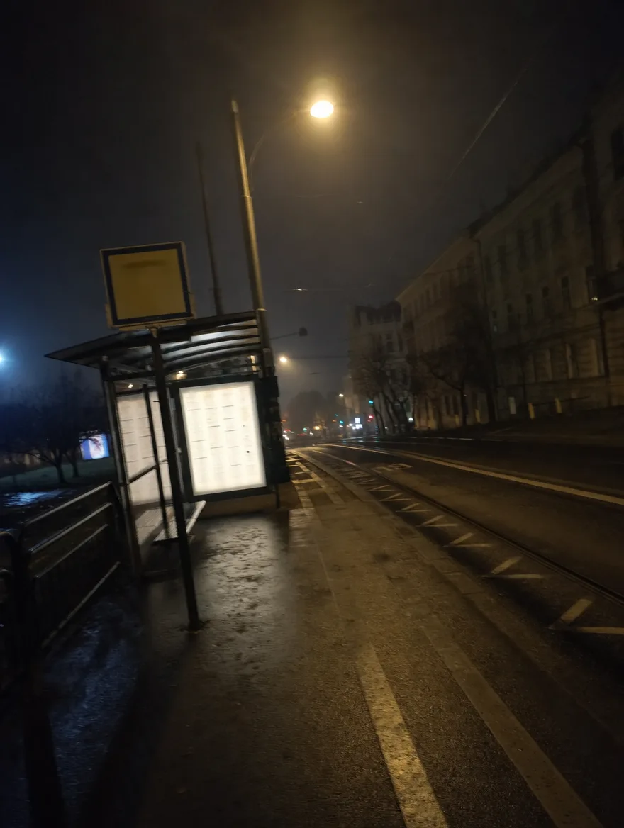 Pusty przystanek nocą, świecący rozkład, mokre tory.